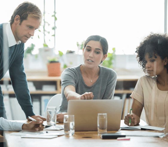 Three colleagues gathered around a laptop in a meeting room, working through a plan together.
